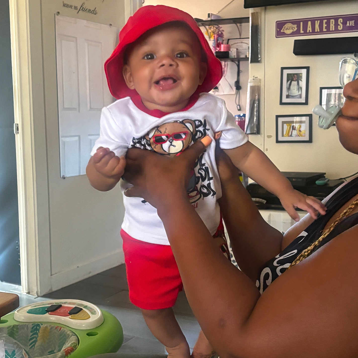 Charles Smith joined the competition — help win amazing prizes! baby, child, smile, red_hat, red_shorts, adult_hand, indoor, living_room, wall_decal, lakers_sign, family_photos, pacifier, joyful, person, holding, furniture, door, ceiling_fan, clothing, portrait