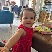 Louise participe au concours pour gagner de l'argent avec cette photo : toddler, child, red_dress, toy, table, living_room, furniture, floor_tile, curtain, window, chair, refrigerator, box, clock, smiling, hair_tie, person, indoor, playful, colorful