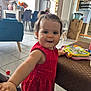 toddler, child, red_dress, toy, table, living_room, furniture, floor_tile, curtain, window, chair, refrigerator, box, clock, smiling, hair_tie, person, indoor, playful, colorful