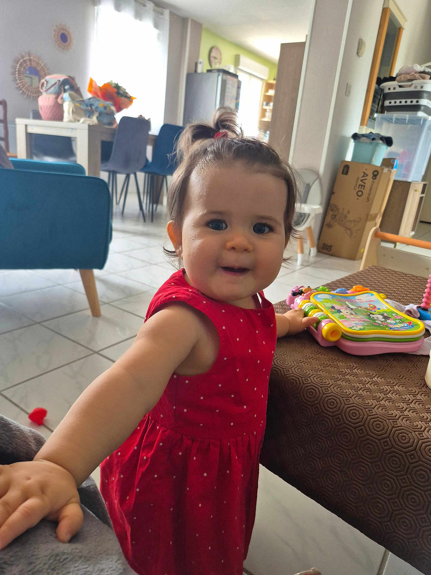 Louise participe au concours pour gagner de l'argent avec cette photo : toddler, child, red_dress, toy, table, living_room, furniture, floor_tile, curtain, window, chair, refrigerator, box, clock, smiling, hair_tie, person, indoor, playful, colorful