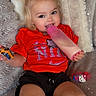toddler, child, portrait, smiling, sitting, bottle, drinking, red_shirt, black_shorts, blonde_hair, pigtails, indoors, blanket, couch, toy, bracelet, cozy, happy, closeup, photography
