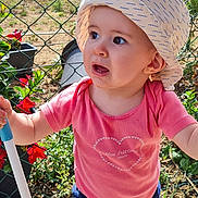 Emy participe au concours pour gagner de l'argent avec cette photo : blue_pants, casual_clothing, child, curious, daylight, expression, fence, flowers, garden, hat, nature, outdoor, pink_shirt, plants, pole, standing, sunlight, toddler, white_shoes, young_child
