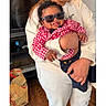 baby, child, person, holding, sunglasses, red_checkered_shirt, navy_pants, indoor, happy, smiling, tongue_out, adult, hand, clothing, floor, wooden_floor, shopping_bag, television, toy_car, casual