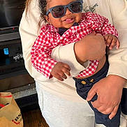 Aaron is registered to the contest to win money with this photo: baby, child, sunglasses, red_shirt, checkered, navy_pants, bow_tie, smiling, tongue_out, indoor, adult, holding, person, wood_floor, bag, casual_clothing, happy, cute, portrait, family