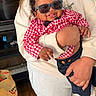 baby, child, sunglasses, red_shirt, checkered, navy_pants, bow_tie, smiling, tongue_out, indoor, adult, holding, person, wood_floor, bag, casual_clothing, happy, cute, portrait, family