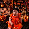 baby, infant, orange_dress, black_headband, pumpkin, jack_o_lantern, lantern, skeleton, halloween, holiday_decor, string_lights, sign, spooky, festive, cute, indoors, celebration, child, costume, holiday