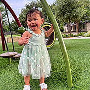 Zoe Sierra is registered to the contest to win money with this photo: toddler, child, playground, dress, green_grass, smiling, happy, outdoor, summer, sandals, nature, trees, play_equipment, curved_pole, flower_pattern, person, cute, young_child, park, fun