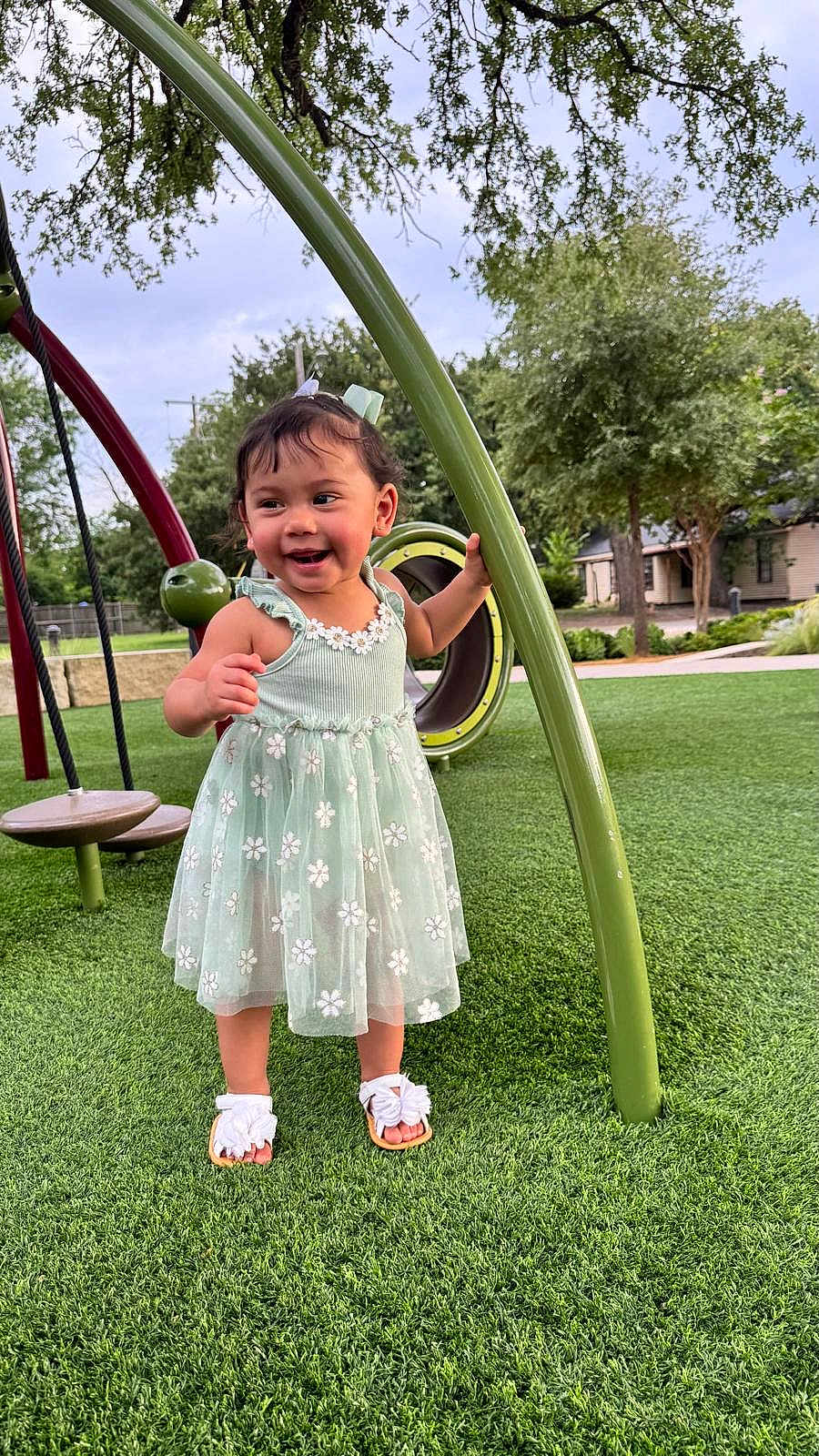 Zoe Sierra is registered to the contest to win money with this photo: toddler, child, playground, dress, green_grass, smiling, happy, outdoor, summer, sandals, nature, trees, play_equipment, curved_pole, flower_pattern, person, cute, young_child, park, fun