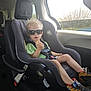 child, toddler, car_seat, sunglasses, curly_hair, shoes, seatbelt, car_interior, window, daylight, casual_clothing, green_shirt, black_car_seat, safety_harness, vehicle, road_trip, cute, confident, young_child, travel