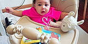 Maria a rejoint le concours — aidez-le/la à gagner de superbes lots ! baby, high_chair, pink_shirt, curly_hair, plush_toy, stuffed_animal, book, indoor, carpet, wooden_floor, furniture, child, person, expression, toy, face, seat, cord, surprise, room
