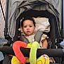 baby, stroller, blanket, soft_toy, colorful, curly_hair, infant, child, seat_belt, fabric, indoors, person, cute, expression, comfort, sitting, playful, closeup, head, portrait