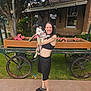 Lilliana May is registered to the contest to win money with this photo: woman, dog, pet, smiling, outdoor, flower_box, wooden_cart, grass, sidewalk, casual_clothing, black_shorts, black_top, slippers, leash, tree, building, window, greenery, happy, summer