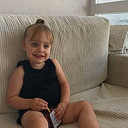 Lahyanna participe au concours pour gagner de l'argent avec cette photo : arm, barefoot, beige, black_clothing, child, container, couch, face, furniture, hair_bun, happy, home, indoor, legs, relaxed, sitting, skin, smiling, snack, toddler