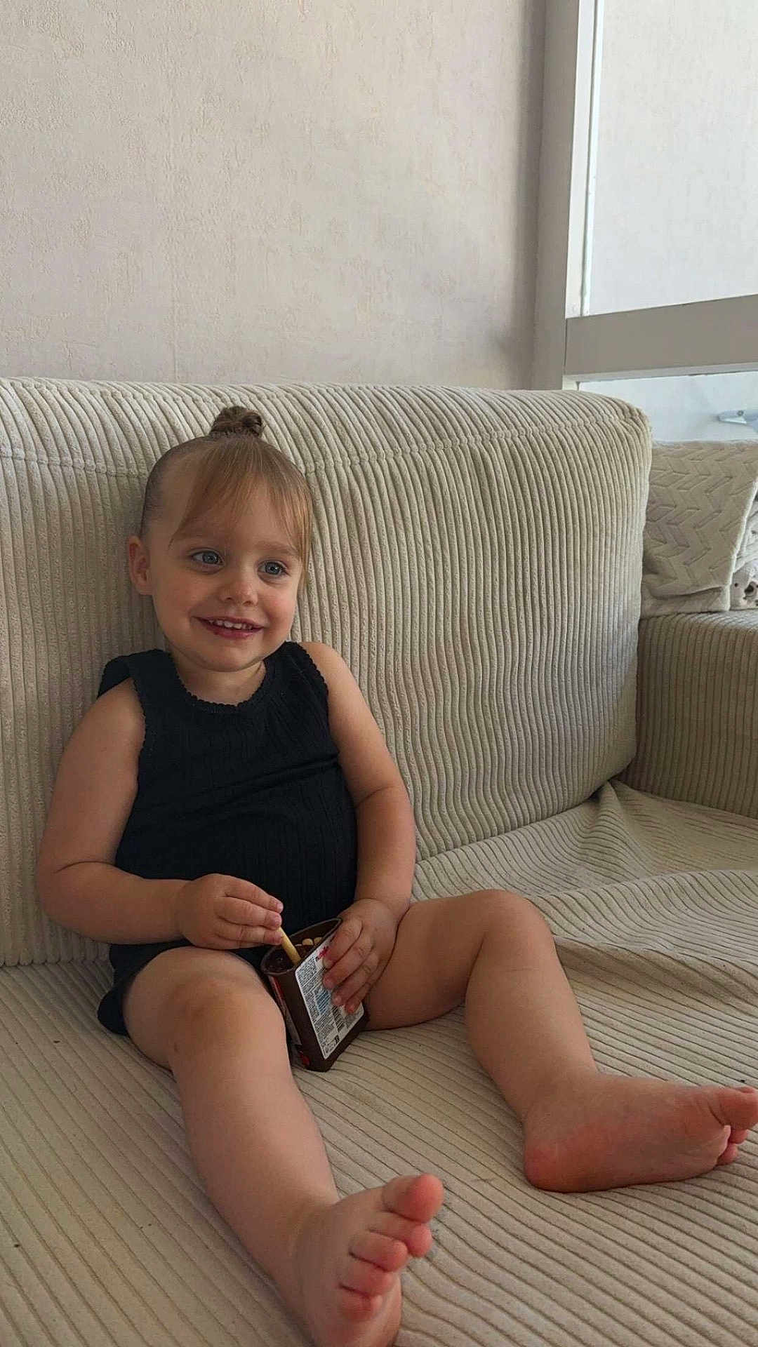 Lahyanna participe au concours pour gagner de l'argent avec cette photo : arm, barefoot, beige, black_clothing, child, container, couch, face, furniture, hair_bun, happy, home, indoor, legs, relaxed, sitting, skin, smiling, snack, toddler