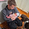 baby, newborn, infant, adult, man, father, bottle_feeding, bottle, blanket, hat, pink_clothing, striped_clothing, dog, spaniel, pet, family, indoor, kitchen, wooden_floor, portrait