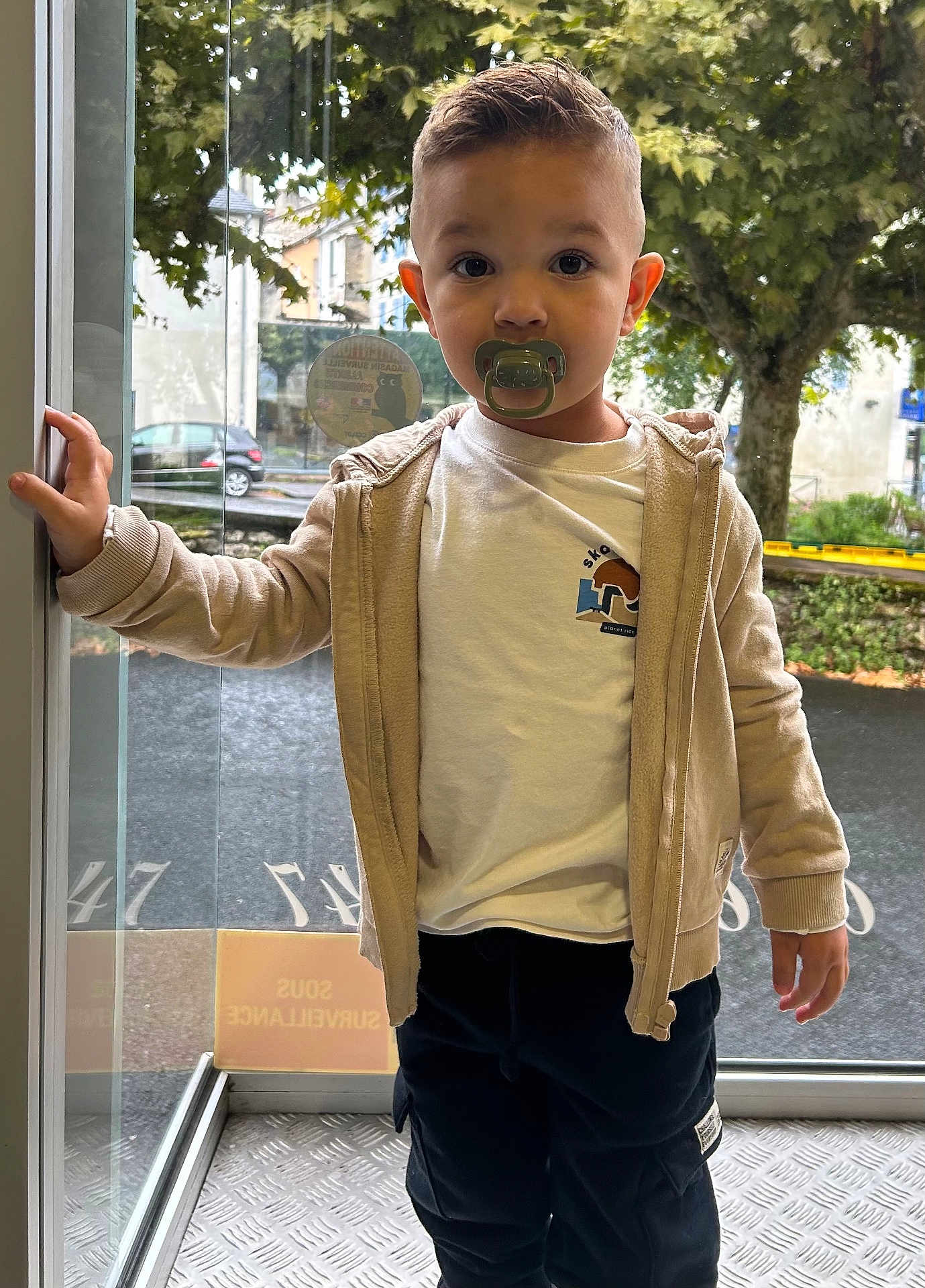 Noamm a rejoint le concours — aidez-le/la à gagner de superbes lots ! child, toddler, pacifier, hoodie, tshirt, pants, standing, glass_door, window, tree, outdoor, street, reflection, hand, face, short_hair, casual_clothing, daylight, young_child, curious