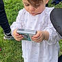 child, toddler, white_dress, grass, outdoors, standing, hands, brown_hair, long_eyelashes, shoes, sneakers, stroller, small_box, candid, portrait, ruffled_sleeves, person, park, curly_wisp, concentration