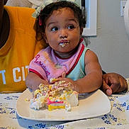Alejandra is registered to the contest to win money with this photo: toddler, child, cake, birthday_cake, messy, food, tablecloth, plate, pink_bib, headband, adult_hand, indoor, celebration, happy, eating, person, young_child, curly_hair, colorful, party