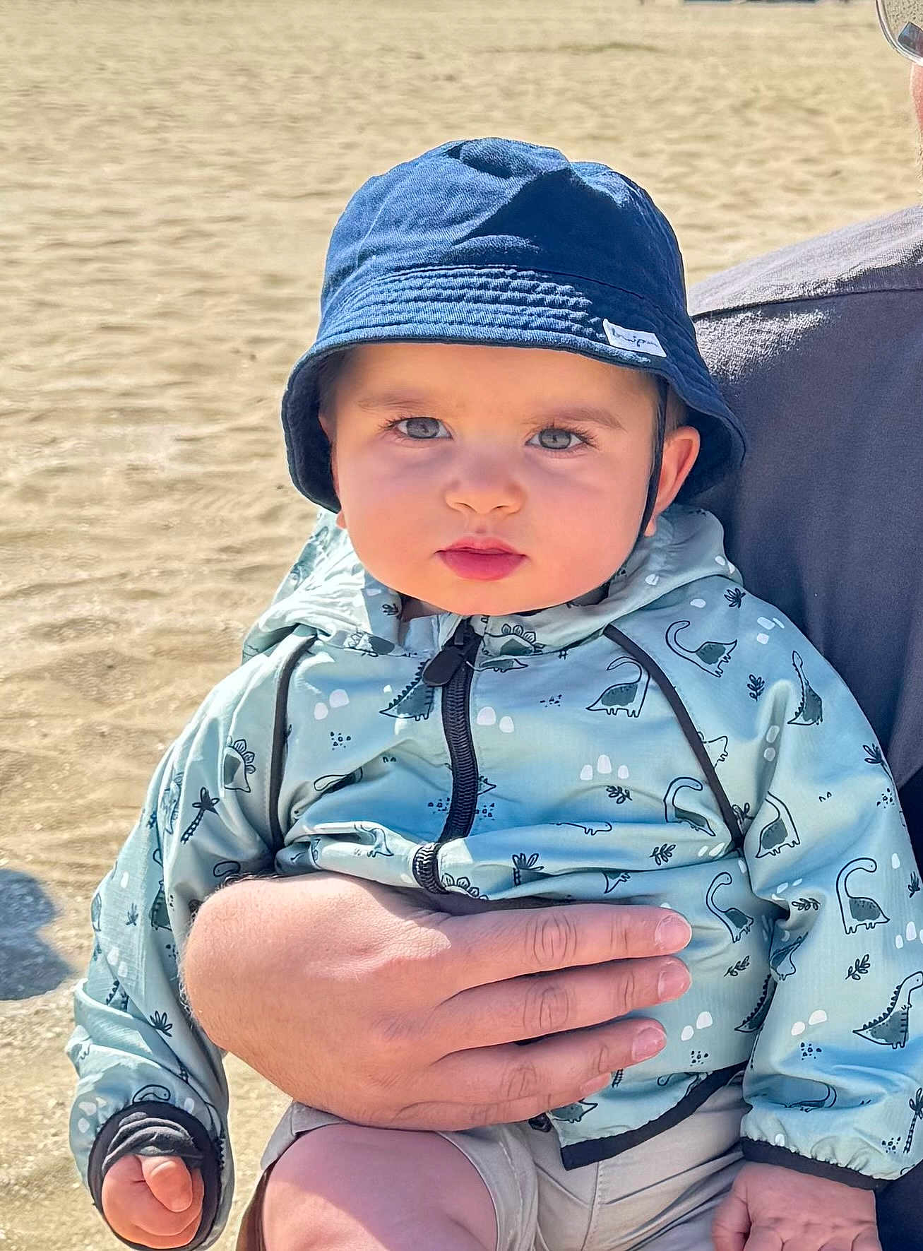 Hussain participe au concours pour gagner de l'argent avec cette photo : toddler, child, baby, hat, bucket_hat, jacket, dinosaur_pattern, hand, beach, sand, sunlight, outdoor, cute, serious_expression, person, clothing, portrait, holding, casual, summer