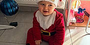 Isaac a rejoint le concours — aidez-le/la à gagner de superbes lots ! toddler, child, christmas, santa_hat, red_clothing, indoor, floor, toy, dollhouse, ornament, holiday, festive, white_tree, decor, smiling, cute, person, home, playful, seasonal