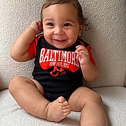 Isaiah is registered to the contest to win money with this photo: baby, baltimore_orioles, barefoot, child, clothing, curly_hair, cute, face, feet, happy, indoor, infant, onesie, person, portrait, sitting, skin, smiling, soft_texture, young_child