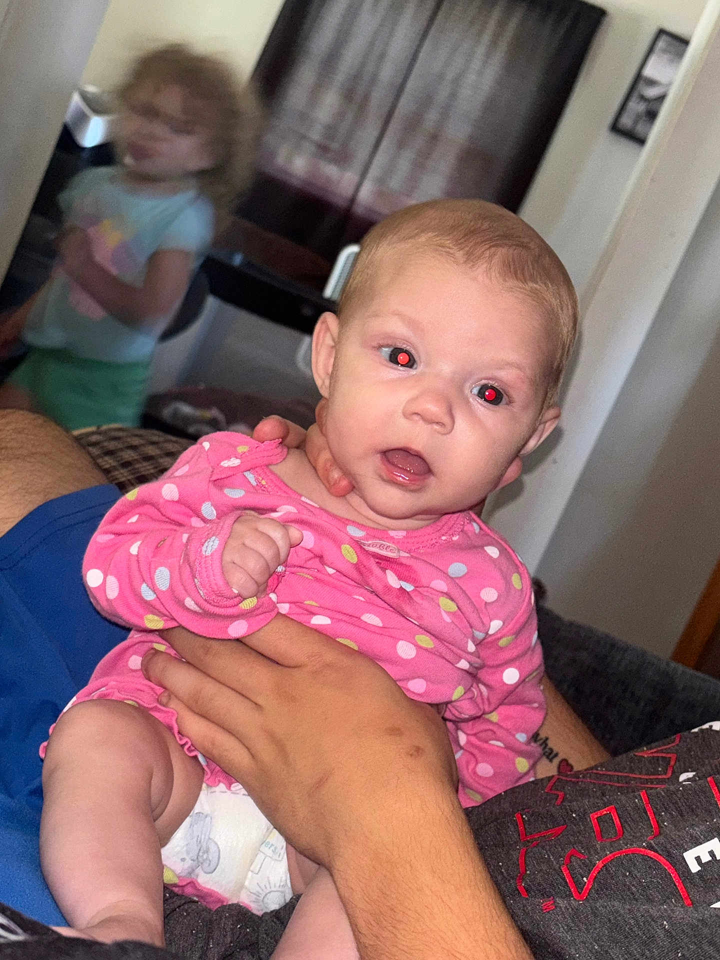 Ava joined the competition — help win amazing prizes! baby, child, toddler, indoor, person, holding, pink_clothing, polka_dot, red_eye, tattoo, hand, blurred_background, curtains, diaper, facial_expression, furniture, adult_hand, shorts, shirt, living_room