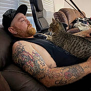Minnie joined the competition — help win amazing prizes! cat, tabby_cat, man, person, beard, tattoos, black_shirt, beanie, indoor, living_room, leather_couch, reclining, sitting, pet, owner, animal, whiskers, window_blinds, massage_chair, cozy