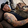 cat, tabby_cat, man, person, beard, tattoos, black_shirt, beanie, indoor, living_room, leather_couch, reclining, sitting, pet, owner, animal, whiskers, window_blinds, massage_chair, cozy