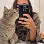 cat, person, selfie, mirror, smartphone, phone_case, ring, sweater, cat_whiskers, cat_paws, fur, indoor, bathroom, shelf, reflection, portrait, hand, table, cozy, pet