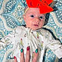 Marlow is registered to the contest to win money with this photo: baby, infant, face, hand, nail_polish, orange_bow, onesie, patterned_blanket, blue, white, red, green, holiday_theme, closeup, person, cute, lying_down, expression, skin, fingers