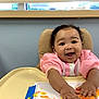 art, baby, blue_wall, child, colorful, curly_hair, expression, face, hands, happy, highchair, indoor, painting, person, pink_clothing, playful, seated, smiling, window, young_child