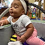 Lovely is registered to the contest to win money with this photo: baby, infant, child, headband, white_shirt, pink_pants, toy, play_area, cushion, sock, indoor, curly_hair, cute, smiling, seated, colorful, soft_toys, carpet, expression, young_child