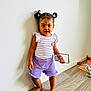 barefoot, casual_clothing, child, cute, girl, home, indoor, person, pigtails, playroom, portrait, purple_shorts, smartphone_case, standing, striped_shirt, toddler, toy, white_wall, wood_floor, young_child