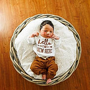 Preston is registered to the contest to win money with this photo: baby, newborn, basket, blanket, wooden_floor, clothing, infant, cute, sleepy, resting, indoors, soft, patterned_blanket, brown_pants, white_shirt, text_on_clothing, small_feet, peaceful, portrait, cozy