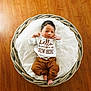 baby, newborn, basket, blanket, wooden_floor, clothing, infant, cute, sleepy, resting, indoors, soft, patterned_blanket, brown_pants, white_shirt, text_on_clothing, small_feet, peaceful, portrait, cozy
