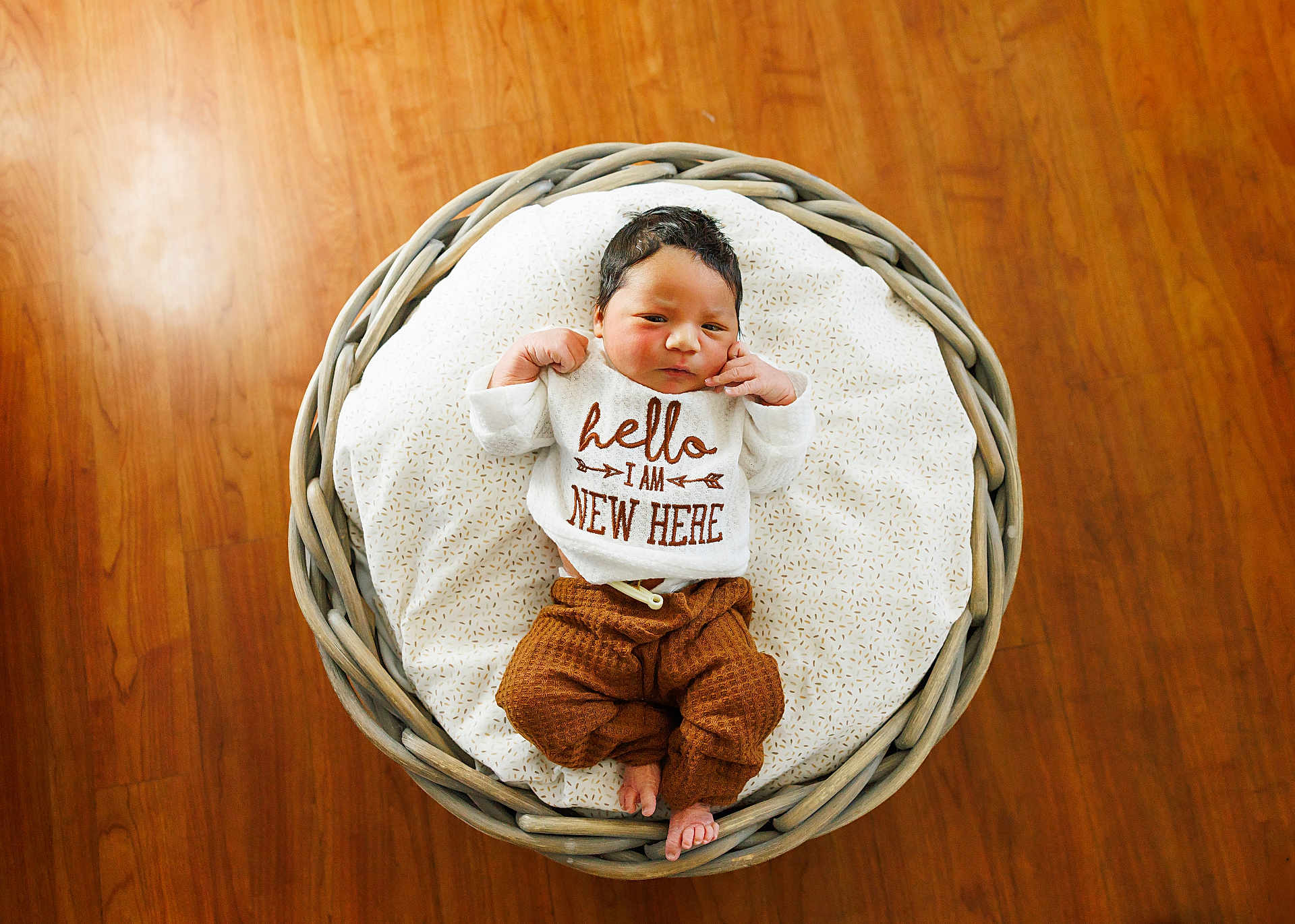 Preston is registered to the contest to win money with this photo: baby, newborn, basket, blanket, wooden_floor, clothing, infant, cute, sleepy, resting, indoors, soft, patterned_blanket, brown_pants, white_shirt, text_on_clothing, small_feet, peaceful, portrait, cozy
