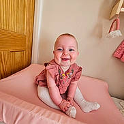 Evelyn is registered to the contest to win money with this photo: baby, blue_eyes, child, chubby_cheeks, clothing, cute, decor, embroidered_outfit, happy, indoors, infant, nursery, person, pink_changing_pad, sitting, smiling, soft_lighting, wall, white_tights, wooden_door