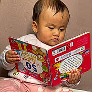 Viona is registered to the contest to win money with this photo: book, child, clothing, cozy, earring, education, expression, face, furniture, hand, indoor, person, pink_pants, portrait, reading, striped_socks, texture, toddler, toy, wall