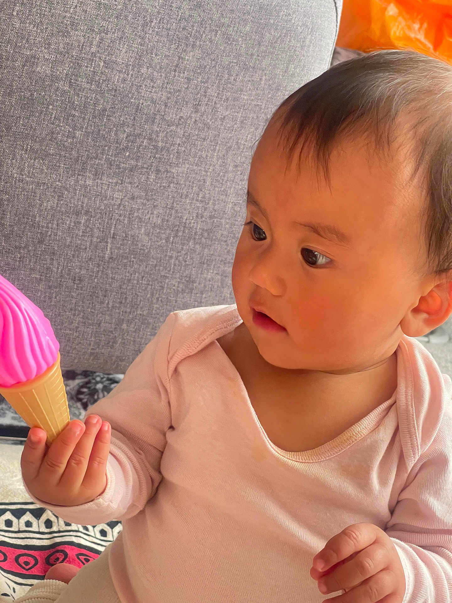 Viona joined the competition — help win amazing prizes! baby, child, toy, ice_cream_cone, pink, curious, couch, patterned_blanket, indoors, infant, clothing, hand, face, looking, sitting, soft_lighting, portrait, cute, young, exploring