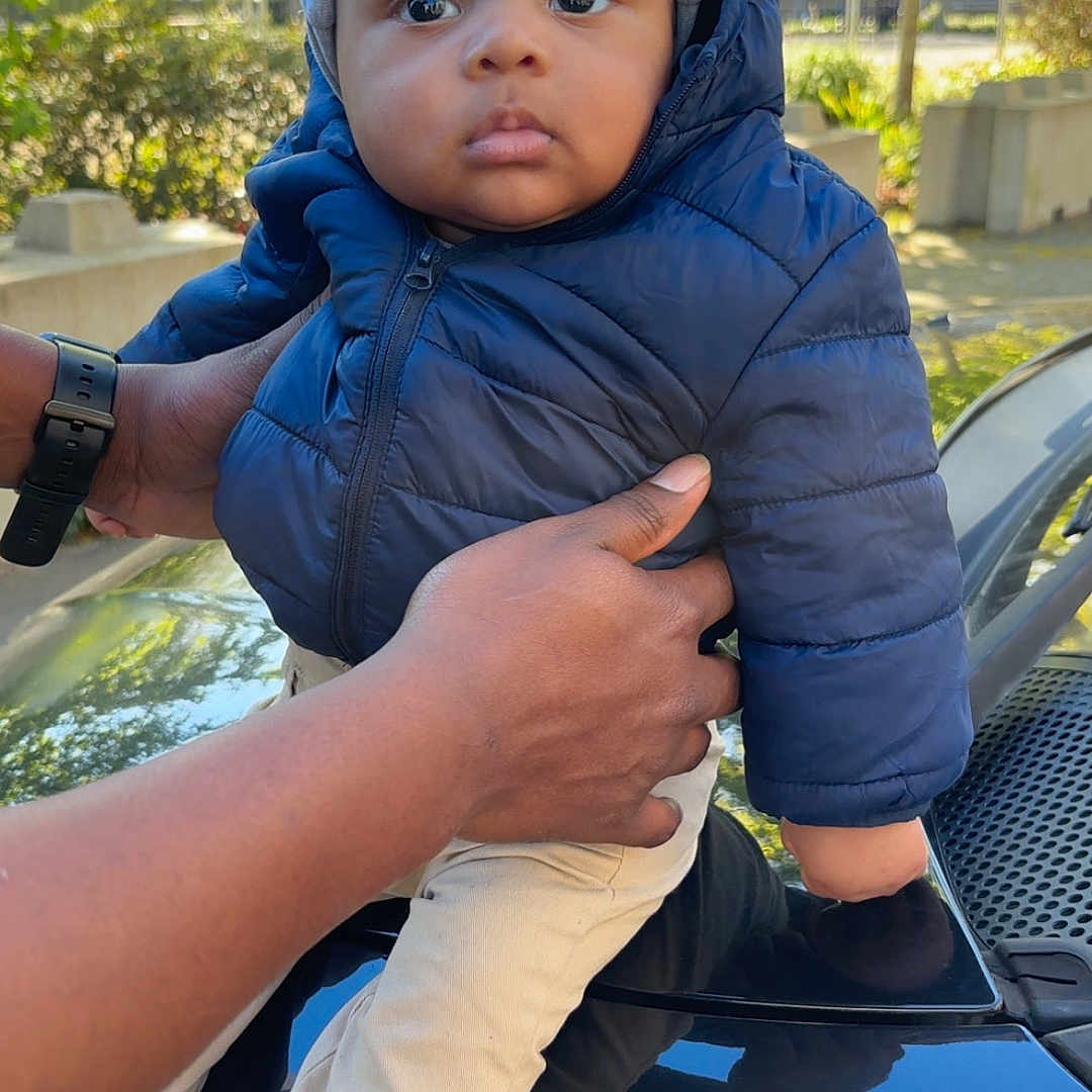 Rayan participe au concours pour gagner de l'argent avec cette photo : baby, child, puffer_jacket, beanie, outdoor, hands, car, reflection, sunlight, trees, greenery, curious, face, portrait, winter_clothing, jacket, pants, sock, person, daylight