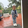 child, boy, smiling, plaid_shirt, jeans, white_sneakers, outdoor, deck, wooden_floor, rain, wet_surface, greenery, trees, string_lights, fence, casual_clothing, happy, standing, daylight, person