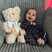 Ares is registered to the contest to win money with this photo: baby, teddy_bear, plush_toy, armchair, checkered_clothing, smiling, happy, infant, cute, cozy, indoor, child, soft_texture, ribbon, portrait, seated, feet, face, furniture, decor