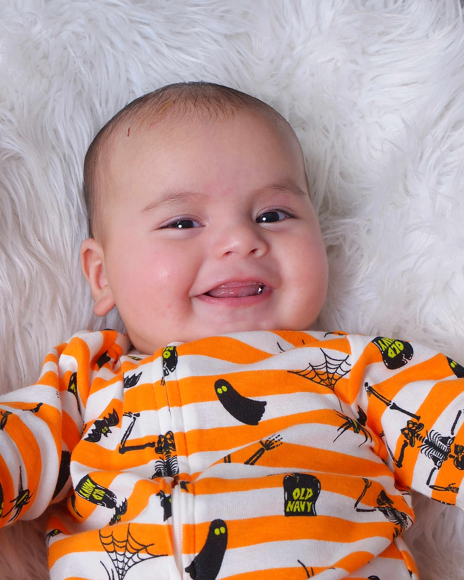 Ares is registered to the contest to win money with this photo: baby, smiling, infant, face, cute, onesie, orange, white, striped_clothing, ghost, skeleton, halloween_theme, tongue_out, soft_blanket, fluffy, close_up, child, happy, portrait, lying_down