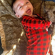 Emriella is registered to the contest to win money with this photo: baby, child, smiling, red_clothing, checkered_pattern, blanket, soft_texture, lying_down, happy, infant, face, portrait, cozy, indoor, warm_lighting, cute, person, young_child, black_and_red, sleepwear