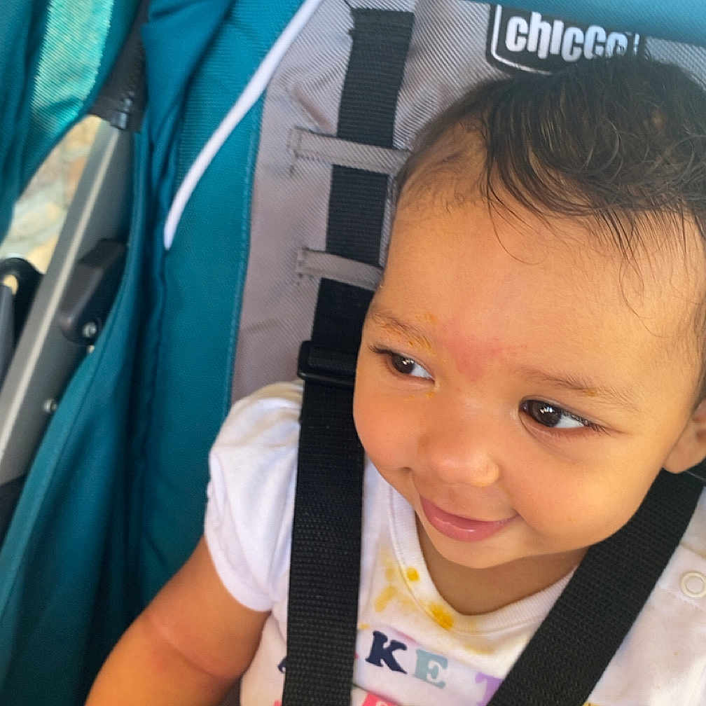 Emriella is registered to the contest to win money with this photo: baby, black_hair, child, closeup, clothing, cute, face, hand, happy, infant, person, portrait, safety_harness, seat, seatbelt, smile, stroller, teal, white_shirt, young