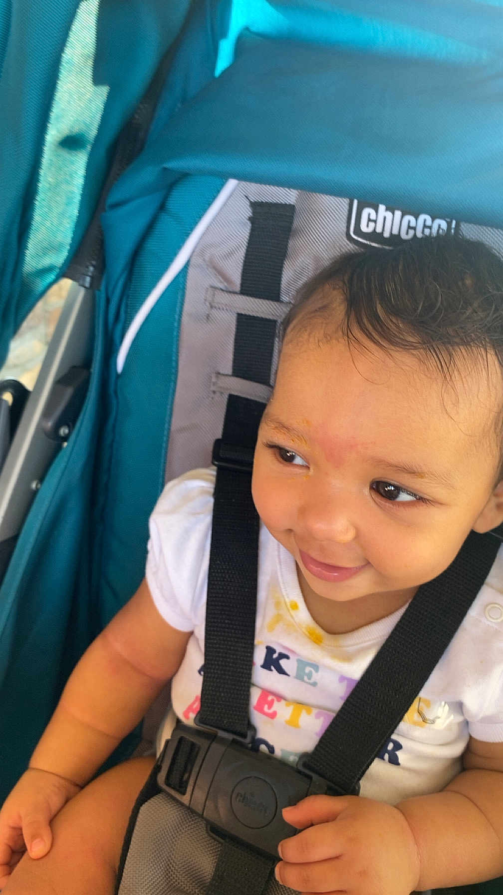 Emriella is registered to the contest to win money with this photo: baby, child, stroller, smile, face, safety_harness, white_shirt, teal, clothing, hand, seat, infant, cute, happy, portrait, young, person, closeup, black_hair, seatbelt