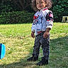 child, toddler, curly_hair, jacket, jeans, shoes, grass, outdoor, sunlight, smile, pose, nature, tree, bushes, playful, casual_clothing, daylight, young_child, happy, person