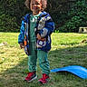 child, smiling, outdoor, grass, jacket, curly_hair, red_shoes, green_pants, sunlight, playground, nature, happy, person, casual_clothing, tree, daylight, toy, lawn, standing, park