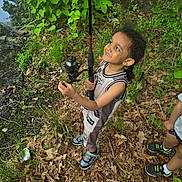Tyrek is registered to the contest to win money with this photo: child, smiling, fishing_rod, outdoor, nature, greenery, leaves, ground, grass, tree, boy, shorts, sneakers, happy, playful, summer, adventure, people, toy, forest