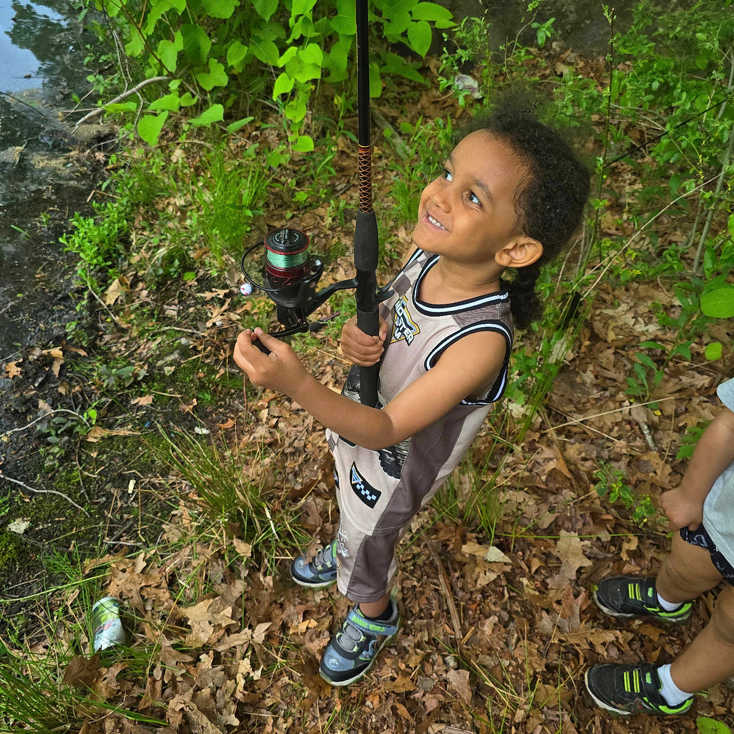 Tyrek is registered to the contest to win money with this photo: child, smiling, fishing_rod, outdoor, nature, greenery, leaves, ground, grass, tree, boy, shorts, sneakers, happy, playful, summer, adventure, people, toy, forest